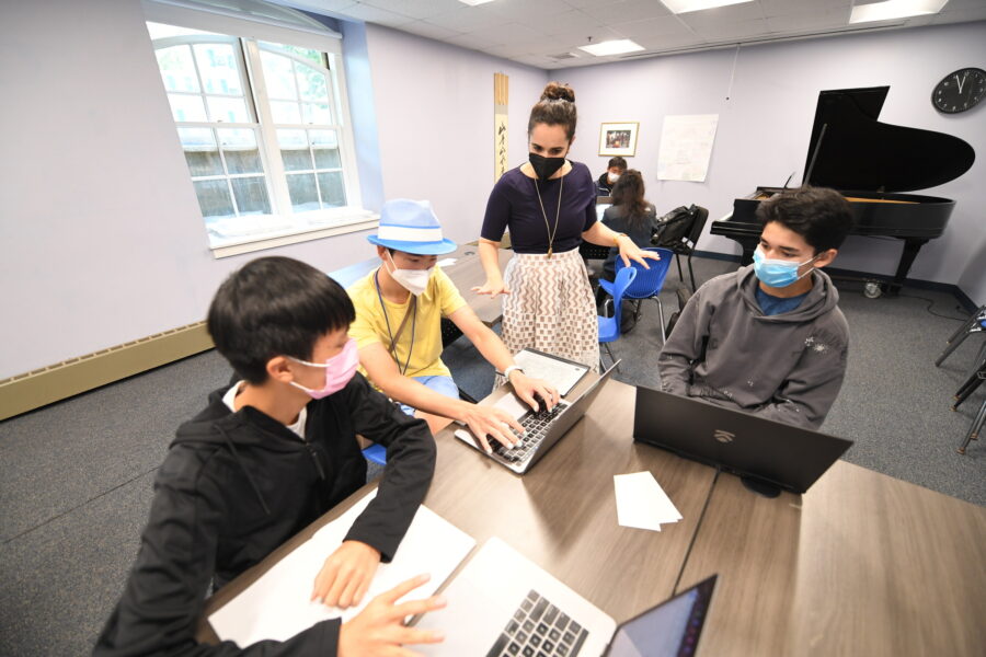 female instructor stands in front of three male students who are seated at a table; they are engaged in conversation